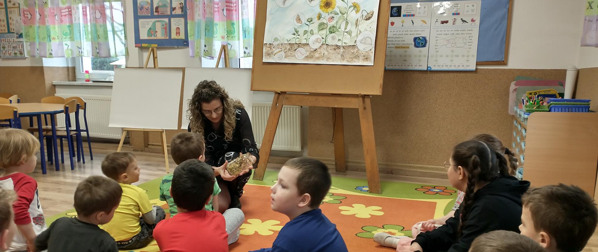 W sali przedszkolnej na kolorowym dywanie w kwiaty siedzą dzieci , którym pani inspektor pokazuje kiełki rzodkiewki umieszczone w słoiku