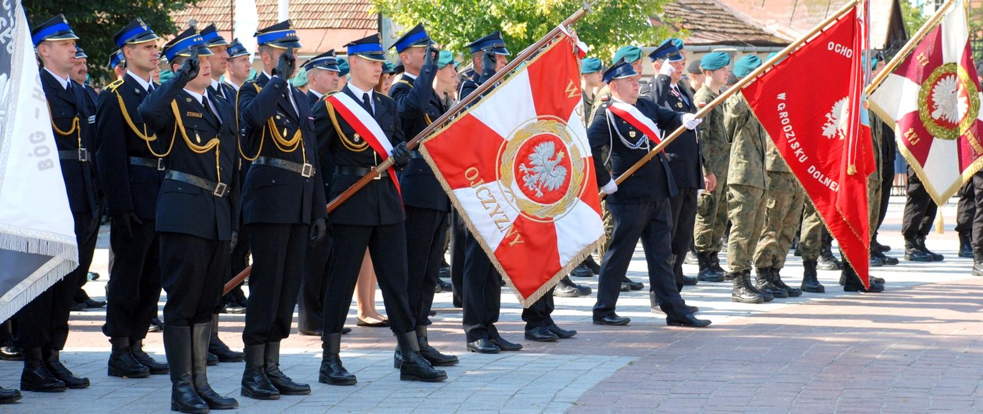 Zdjęcie zrobione na placu. Na zdjęciu widać umundurowanych mężczyzn wśród nich strażacy są to poczty sztandarowe formacji mundurowych część strażaków ma uniesioną prawą dłoń w salucie. Sztandary pocztów są pochylone. 