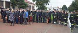 Jubileusz 95-lecia OSP Konary. Plac przed remizą strażacką. Uczestnicy uroczystości w postawie stojącej w trakcie hymnu państwowego. Strażacy w mundurach salutują. W tle budynki i drzewa.