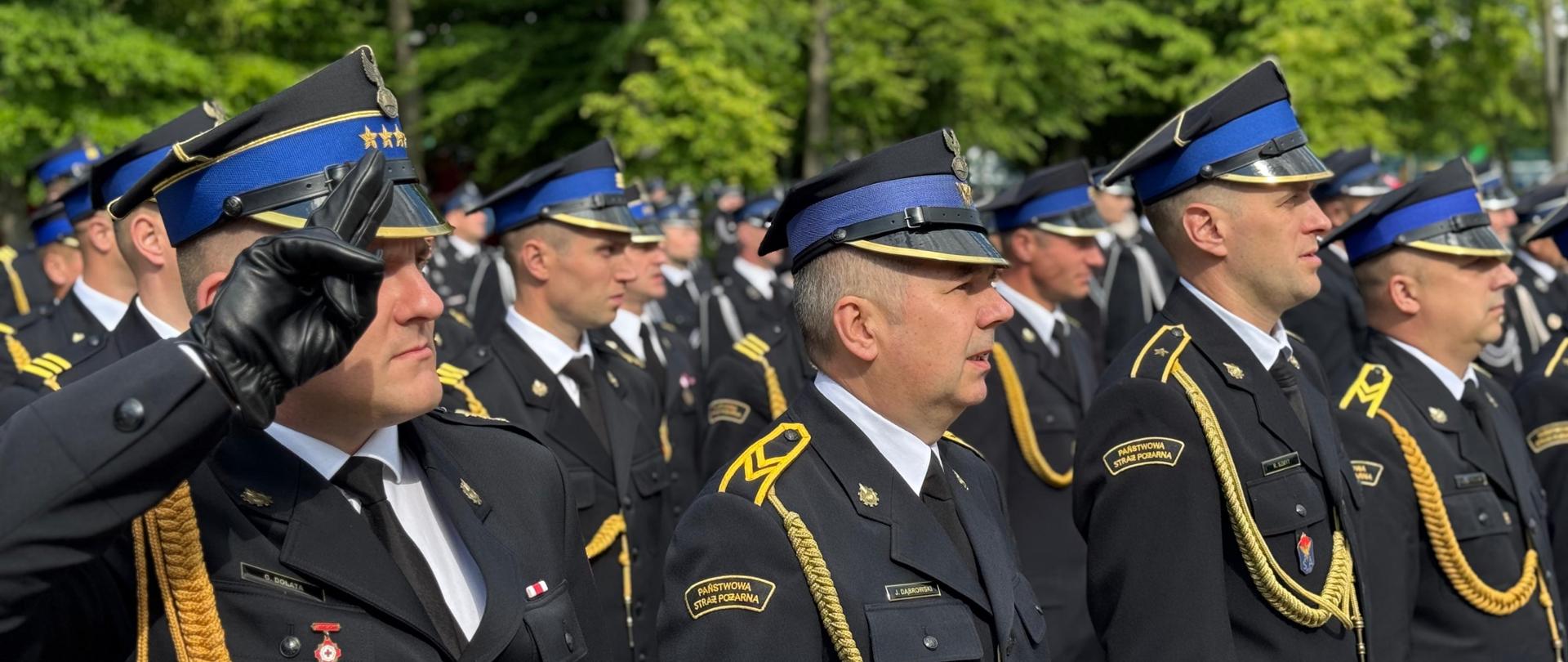 Na zdjęciu widoczni strażacy ubrani w mundury wyjściowe ze sznurem stojący podczas uroczystego apelu. Dowódca pododdziału oddaje honor pozostali strażacy stoją na baczność