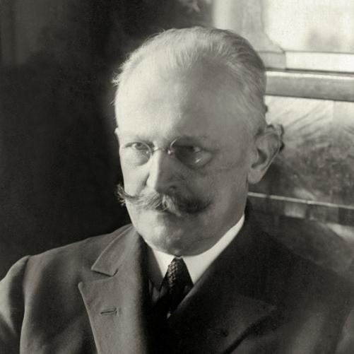 Stanisław Patek - minister spraw zagranicznych. Czarno-białe zdjęcie mężczyzny w okularach i wąsami, w ciemnej dwurzędowej marynarce, ciemny krawat.