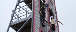 Na zdjęciu widać dwóch uczestników zawodów, którzy wspinają się z drugiego piętra na trzecie piętro wspinalni. W tle niebo pochmurne.