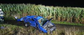 Pora nocna, zdeformowana karoseria niebieskiego samochodu osobowego stojącego na ścieżce rowerowej. Teren przed samochodem mocno zdeformowany, za samochodem pole kukurydzy, z lewej strony zdjęcia strażak w ubraniu specjalnym koloru czarnego z odblaskowymi pionowymi i poziomymi pasami na głowie biały hełm. 