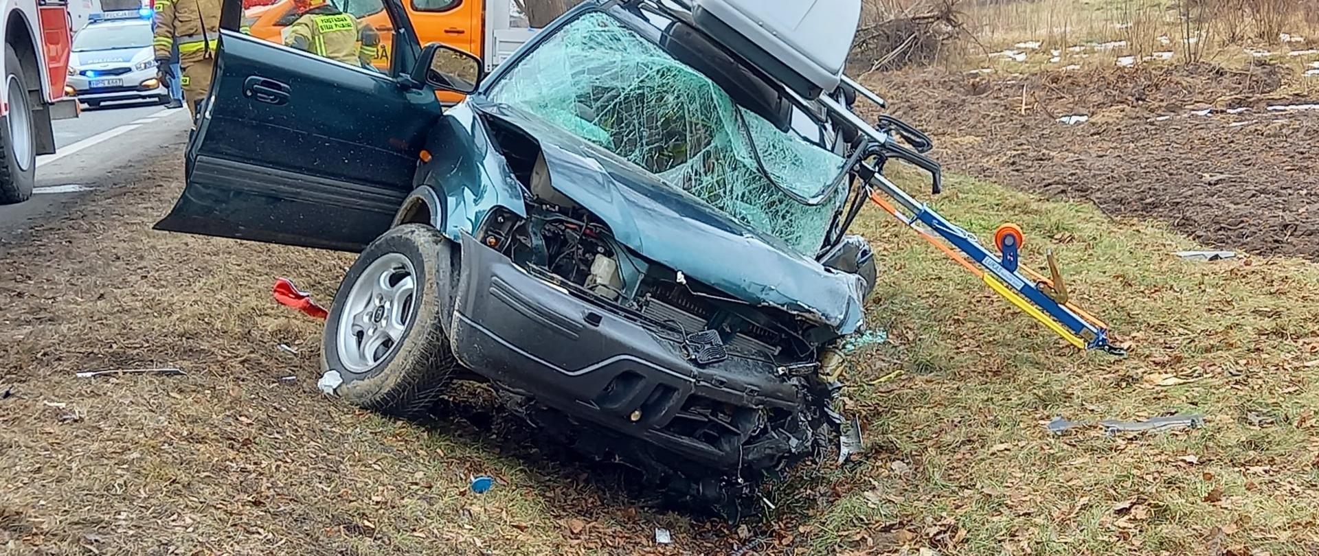 Zdjęcie przedstawia uszkodzony samochód znajdujący się w rowie drogowym. W tle widać samochody Państwowej Straży Pożarnej, Policji, Pomocy Drogowej oraz drzewa.