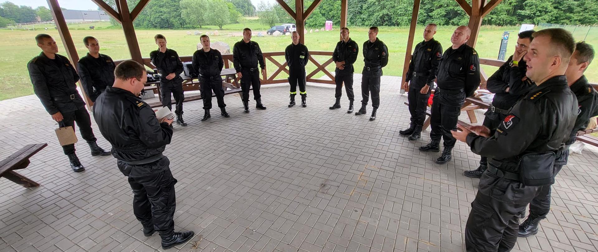 grupa strażaków w czarnych mundurach stoi w kręgu pod drewnianą wiatą, w tle łąka 