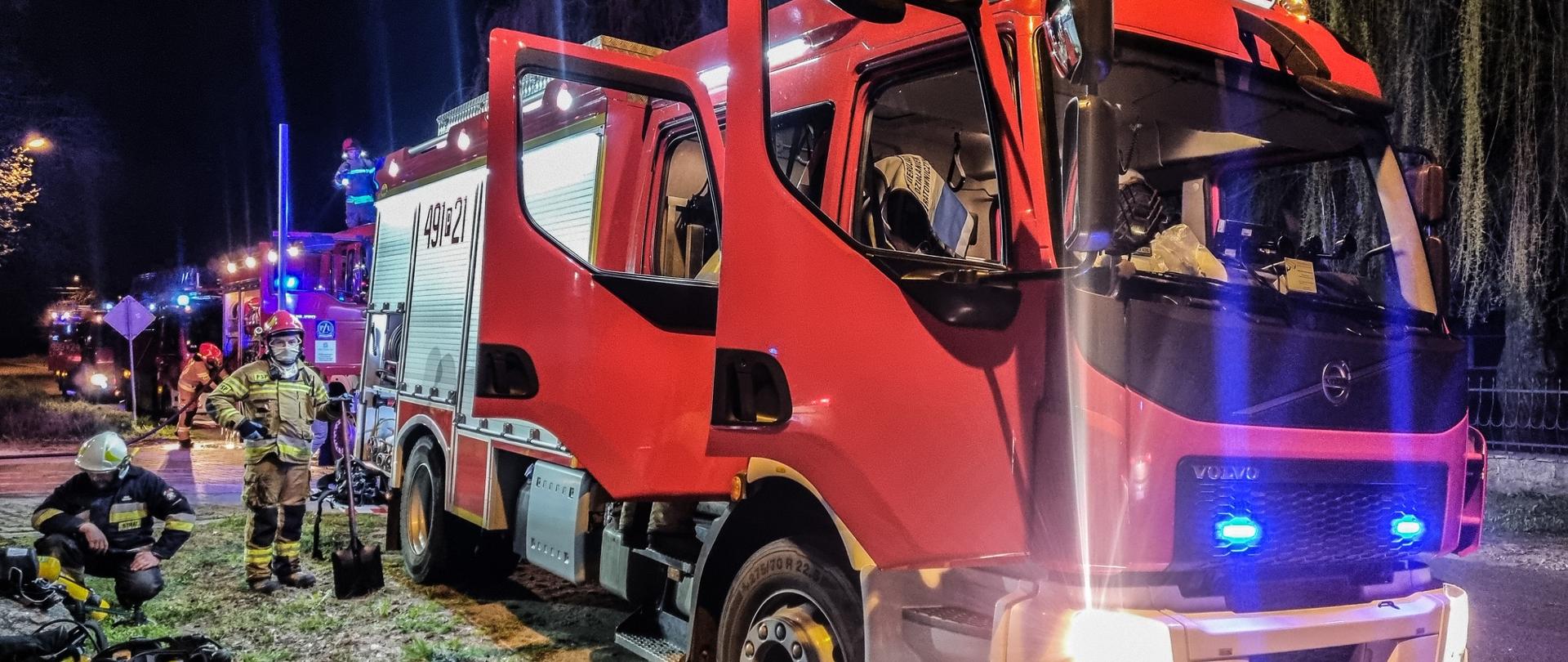 Zdjęcie przedstawia samochód pożarniczy koloru czerwonego, przy samochodzie stoją strażacy, którzy realizowali zadania służbowe podczas gaszenia pożaru pomieszczenia magazynowego.