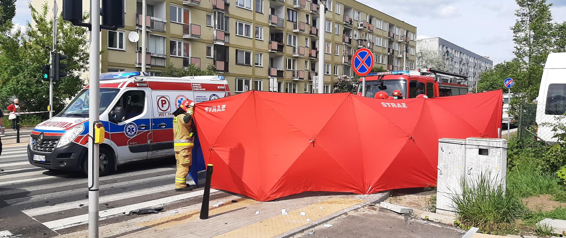Na zdjęciu widać rozstawiony czerwony parawan osłaniający oraz strażaka w piaskowym ubraniu ochronnym. Na pasach stoi biało-czerwono-niebieska karetka Pogotowia ratunkowego .Na drugim planie widać również czerwony samochód straży pożarnej.
