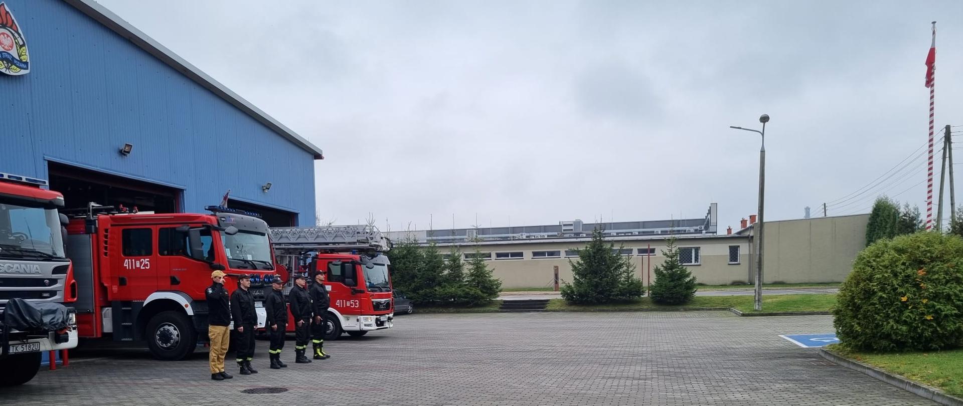 Zdjęcie obrazuje strażaków w umundurowaniu koszarowym. Strażacy znajdują się na tle samochodów ratowniczo-gaśniczych. Po prawej stronie widać maszt, na którym zawieszona jest biało-czerwona flaga.