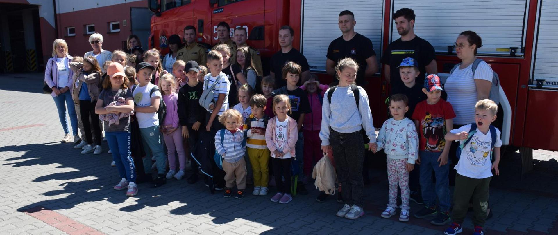 Zdjęcie przedstawia grupę dzieci wraz z opiekunami i funkcjonariuszami PSP na tle wozu ratowniczo-gaśniczego.