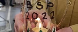 Zdjęcie przedstawia szklany lampion ze świeczką w środku, z napisem BŚP 2022. Lampion trzymany w rękach przez strażaka. 