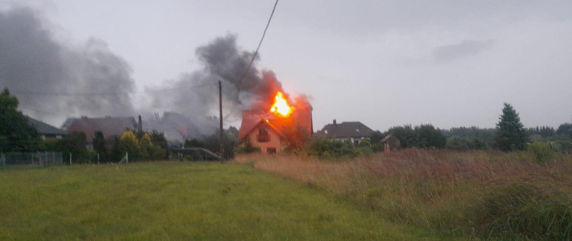 Dzień. Widok na budynek jednorodzinny. Na dachu budynku duże płomienie ognia.