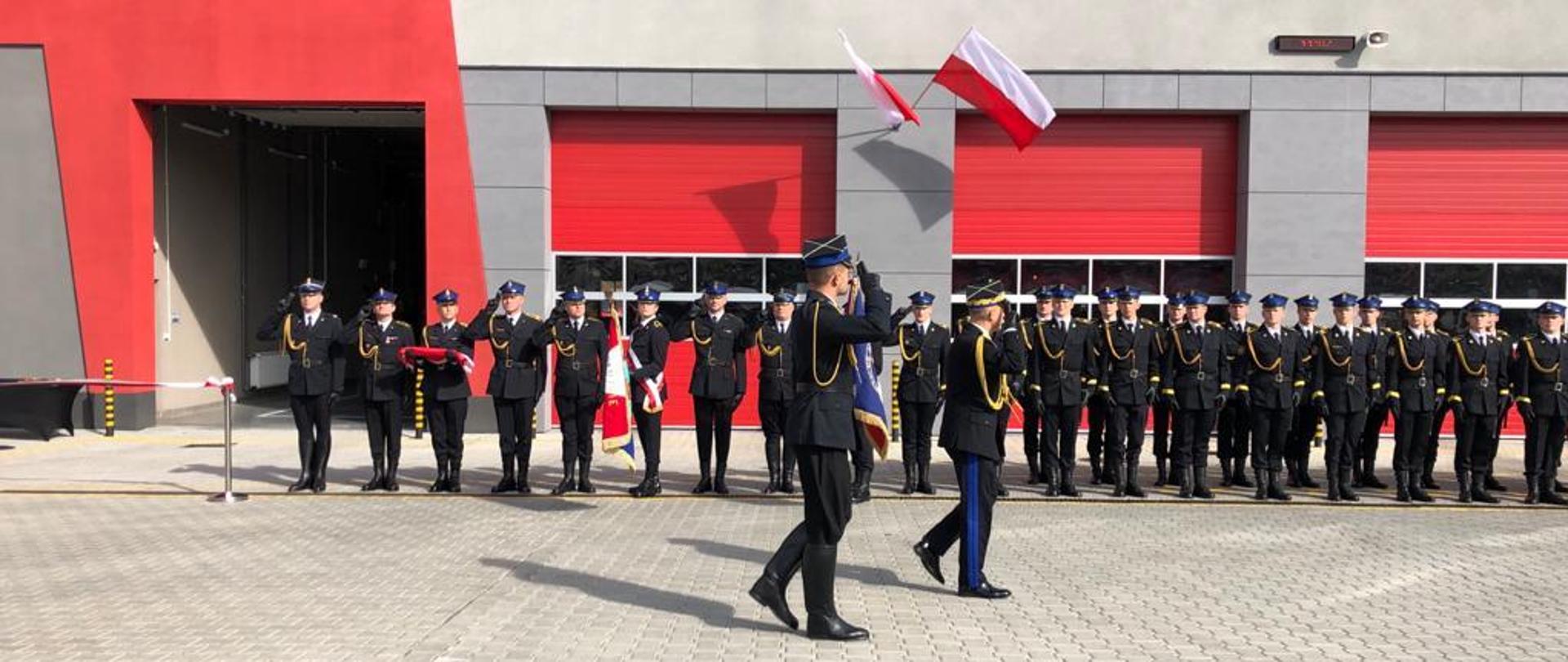 Komendant Główny PSP maszeruje przed pododdziałem strażaków, w tle budynek strażnicy