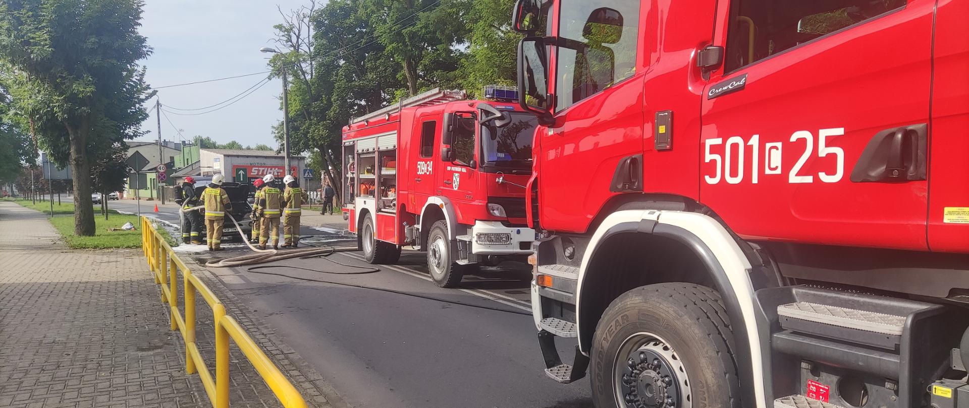Zdjęcie przedstawia drogę wojewódzką, na której znajdują się pojazdy służb ratowniczych (dwa wozy straży pożarnej) strażaków podczas gaszenia pojazdu osobowego.