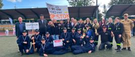 Grupowe zdjęcie uśmiechniętych drużyn strażackich OSP Radwan oraz OSP Stok, pozujących na boisku sportowym po VII wojewódzkich zawodach sportowo-pożarnicze Ochotniczych Straży Pożarnych z terenu województwa łódzkiego. Kobiety oraz mężczyźni, którzy brali udział w zawodach ubrani są w ciemne mundury strażackie, na szyjach mają zawieszone medale. Strażacy trzymają w rękach puchary za osiągnięte miejsce, dyplomy oraz czek z nagroda pieniężną. Z prawej strony grupy stoi Komendant Powiatowy PSP w Opocznie, natomiast z lewej strony prezesi drużyn oraz Pani Burmistrz gminy Białaczów. Cała grupa pozuje z uśmiechem w świetle dziennym n a tle infrastruktury sportowej oraz drzew.