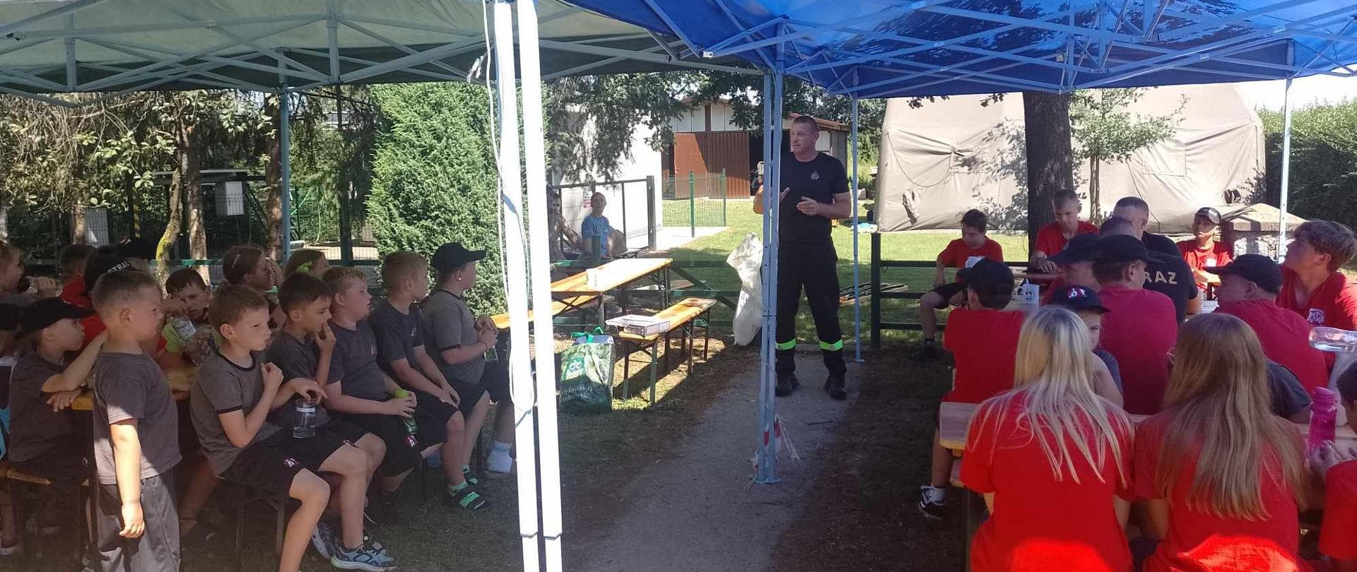 Strażak udziela rad dzieciom, wypoczywającym na obozie harcerskim. Dzieci skupione pod namiotami słuchają prelekcji strażaka. Głównym celem spotkania jest podniesienie bezpieczeństwa najmłodszych w czasie letniego wypoczynku.
