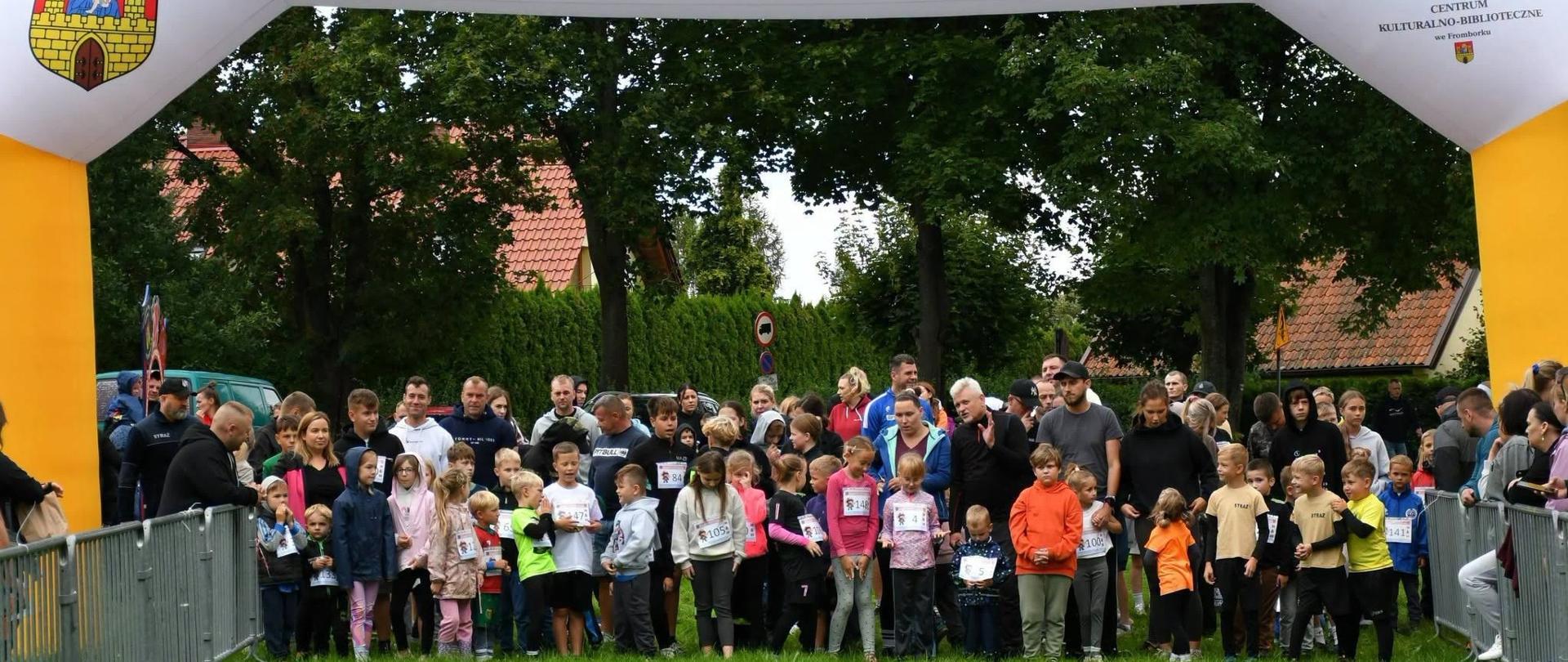 Na zdjęciu grupa dzieci przygotowuje się do startu w biegu pod bramą z napisem „Frombork – Tu zatrzymało się Słońce”. Młodzi uczestnicy, ubrani w kolorowe stroje i numery startowe, stoją na trawie w otoczeniu dorosłych. Trasę wyznaczają metalowe barierki, a w tle widać drzewa i zabudowania.