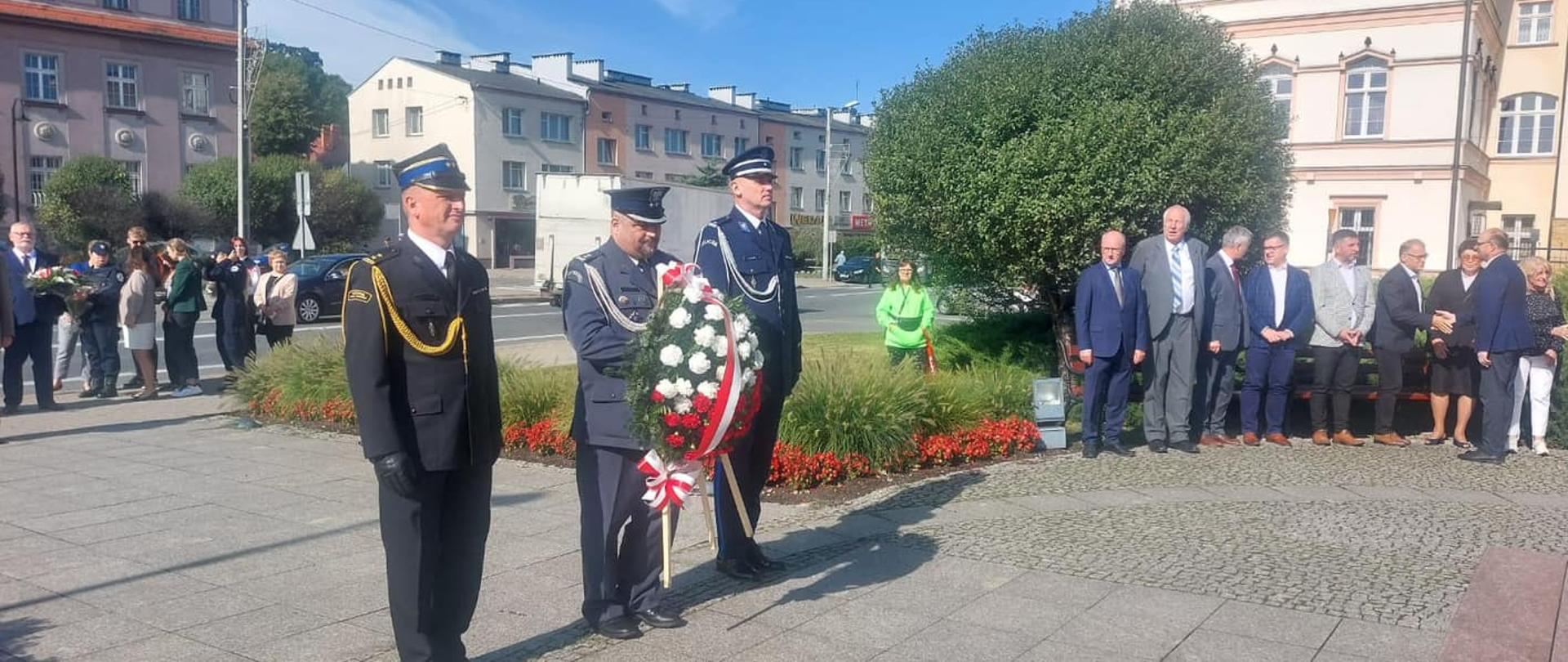 Na zdjęciu znajduje się delegacja składająca kwiaty pod pomnikiem, w skład delegacji wchodzą strażak, policjant i przedstawiciel służby więziennej w mundurach, w tle widać inne osoby oraz budynki