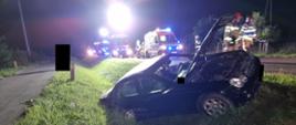 Widać samochód w rowie, jest ciemno, za nim pojazdy straży pożarnej i karetka