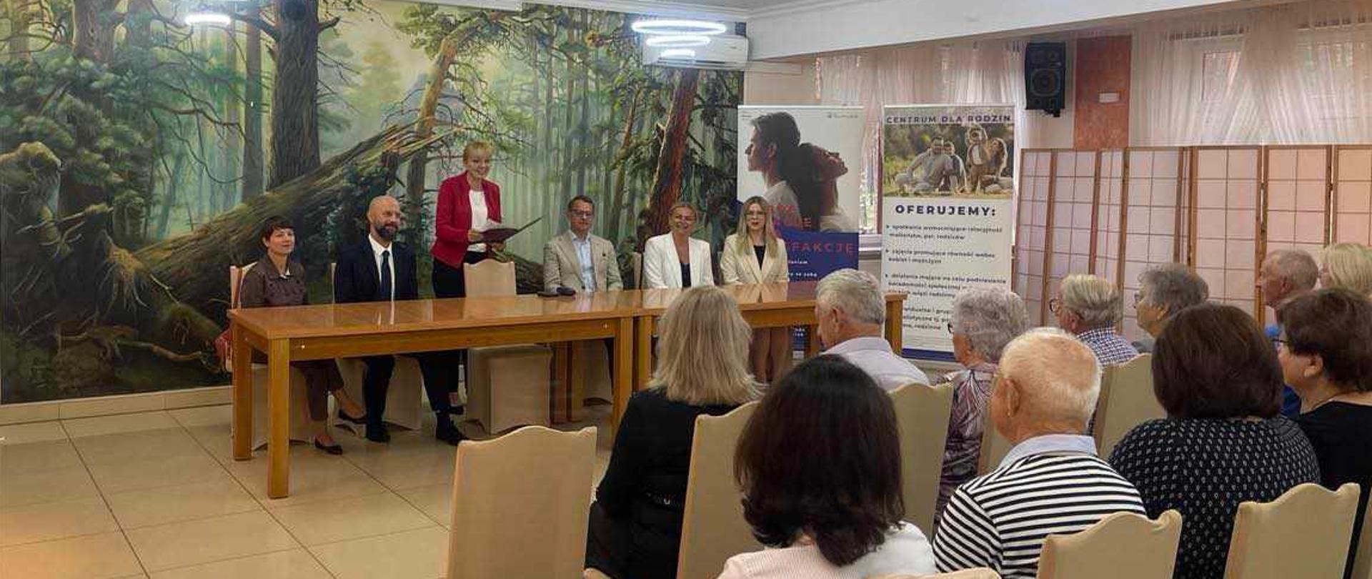 Na pierwszym planie widać osoby siedzące tyłem. Na drugim można dostrzec stół, przy którym znajduje się sześć osób. 
