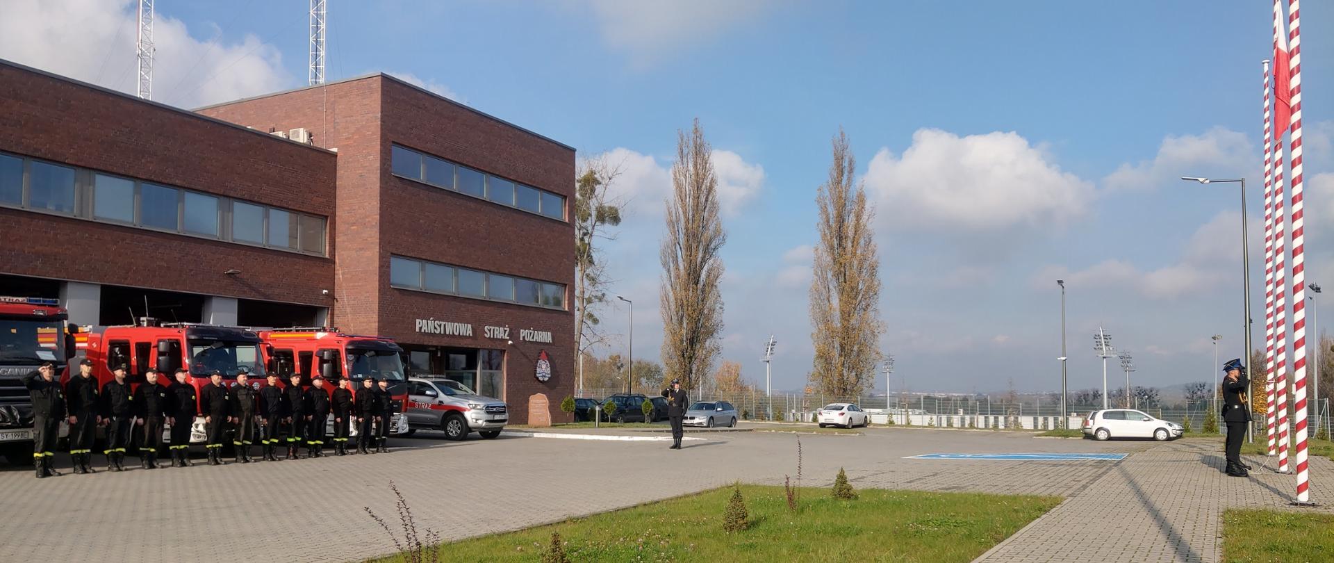Po lewej stronie,w bramach garaży komendy Państwowej Straży Pożarnej stoją samochody ratowniczo - gaśnicze. Na budynku nad wejściem napis Państwowa Straż Pożarna i logo PSP. Przed samochodami stoją w szeregu strażacy w czarnych ubraniach koszarowych. Naprzeciw nich stoją trzy maszty, na środkowym flaga państwowa. Pzred maszatami stoi trzech strażaków w mundurach i rogatywkach. Na środku placu stoi strażak w mundurze, który salutuje. Za nim zaparkowane samochody i płot z siatki. Pomiędzy masztem a garażami trawnik.
