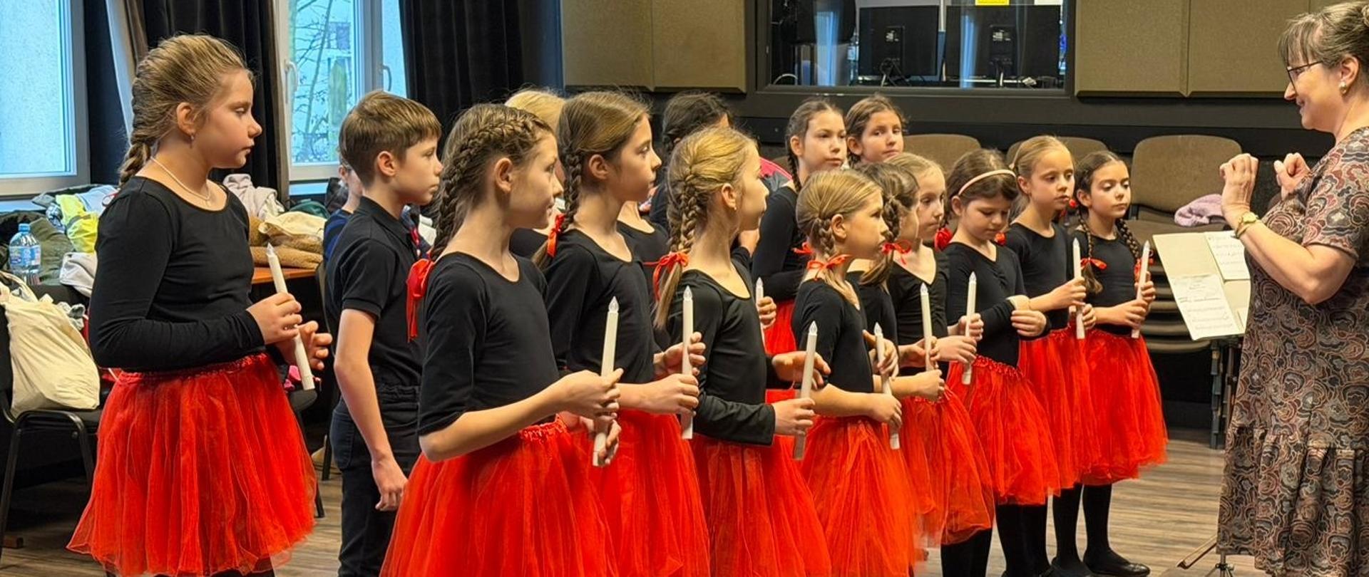 Grupa dzieci ubranych na czarno-czerwono stojąca w sali.