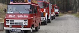 Na zdjęciu widoczna jest kolumna samochodów pożarniczych ustawionych wzdłuż drogi prowadzącej przez teren leśny. W skład zestawienia wchodzą zarówno starsze, jak i nowsze pojazdy jednostek OSP, które przybyły na ćwiczenia „Kozieławy 2025”. Konwój ukazuje różnorodność wyposażenia jednostek biorących udział w działaniach szkoleniowych.