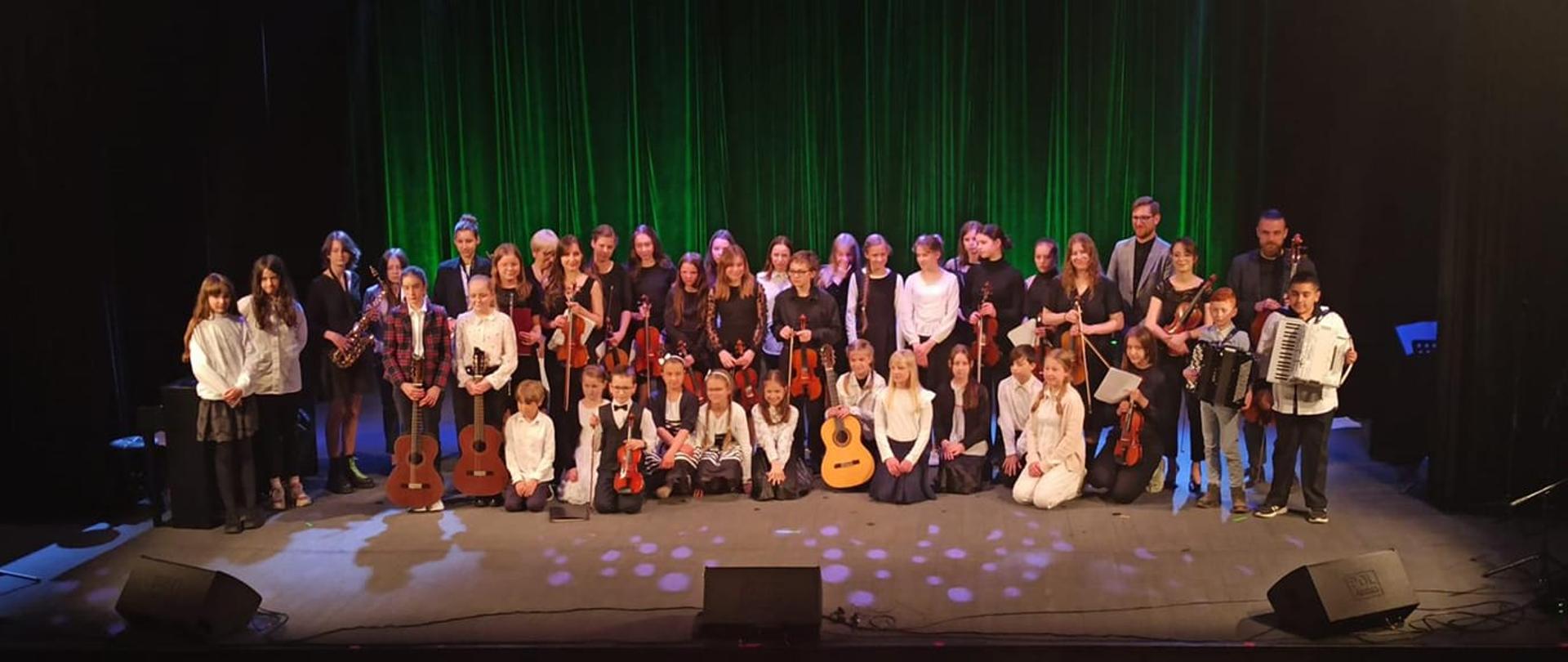 Grupa dzieci trzymających instrumenty wraz z nauczycielami pozujący do zdjęcia grupowego uczestniczące w Koncercie młodych muzyków dla sympatyków szkoły 16 maja 2023, który odbył się w Kłodzkim Ośrodku Kulltury