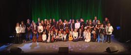 Grupa dzieci trzymających instrumenty wraz z nauczycielami pozujący do zdjęcia grupowego uczestniczące w Koncercie młodych muzyków dla sympatyków szkoły 16 maja 2023, który odbył się w Kłodzkim Ośrodku Kulltury