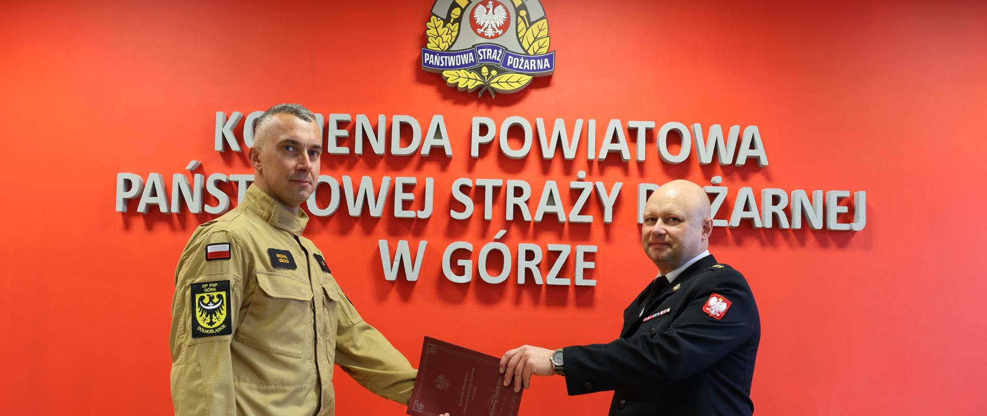 Dwóch funkcjonariuszy Państwowej Straży Pożarnej stoi przed czerwoną ścianą z emblematem PSP i napisem „Komenda Powiatowa Państwowej Straży Pożarnej w Górze”. Funkcjonariusz w mundurze specjalnym wręcza drugiemu dokument w bordowej teczce.