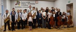 W auli PSM widzimy grupę uśmiechniętych uczniów, trzymających instrumenty muzyczne – wiolonczele i saksofony. Przeważa kolorystyka beżowo – biała.