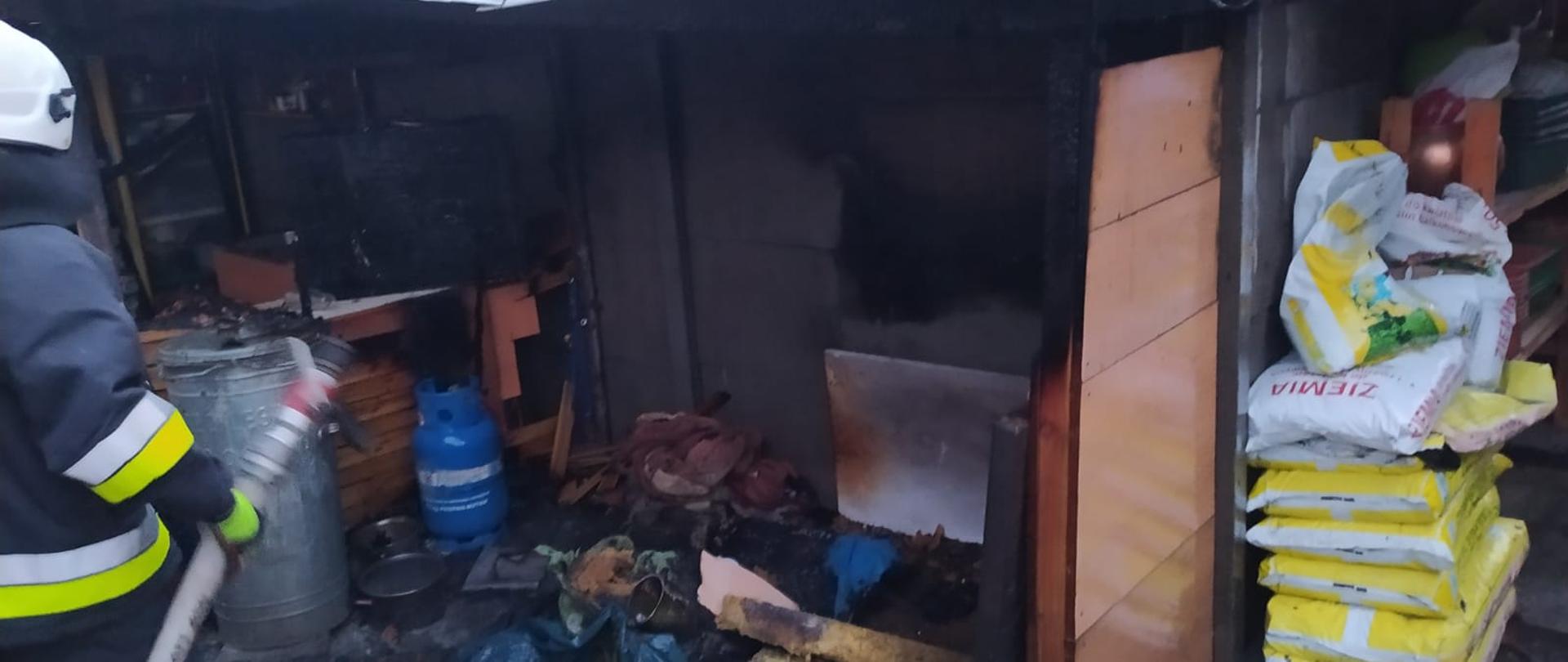 Nadpalone pomieszczenie wraz z resztkami po pożarowymi na ziemi. Obok strażak z wężem. 