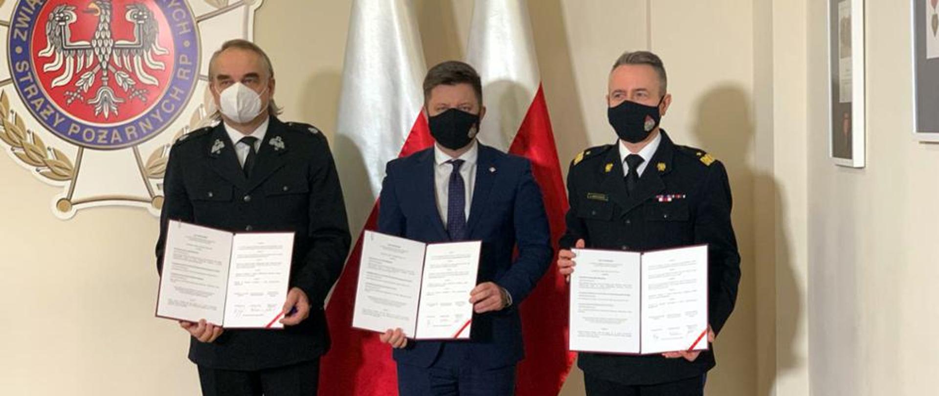 Zdjęcie przedstawia Pana Michała Dworczyk, reprezentującego Kancelarię Prezesa Rady Ministrów, ubranego w garnitur, Komendanta Głównego Państwowej Straży Pożarnej nadbryg. mgr Andrzeja Bartkowiaka oraz Prezesa ZG ZOSP RP Pana Waldemara Pawlaka. Wszyscy trzymają podpisane listy intencyjne. W tle flagi Polski oraz logo ZOSP RP.