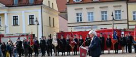 Na pierwszym planie, z prawej strony kadru, widać starszego mężczyznę o siwych, puszystych włosach, ubranego w ciemną marynarkę i jeansy, który stoi przy nowoczesnej, czerwonej mównicy z logo Państwowej Straży Pożarnej. W tle, naprzeciwko przemawiającego, ustawione są poczty sztandarowe ze zdobionymi sztandarami oraz szeregi strażaków w czarnych mundurach galowych. Za nimi rozciąga się duża czerwona ścianka promocyjna, a w oddali widoczne są zabytkowe kamienice oraz grupa mieszkańców obserwujących wydarzenie.