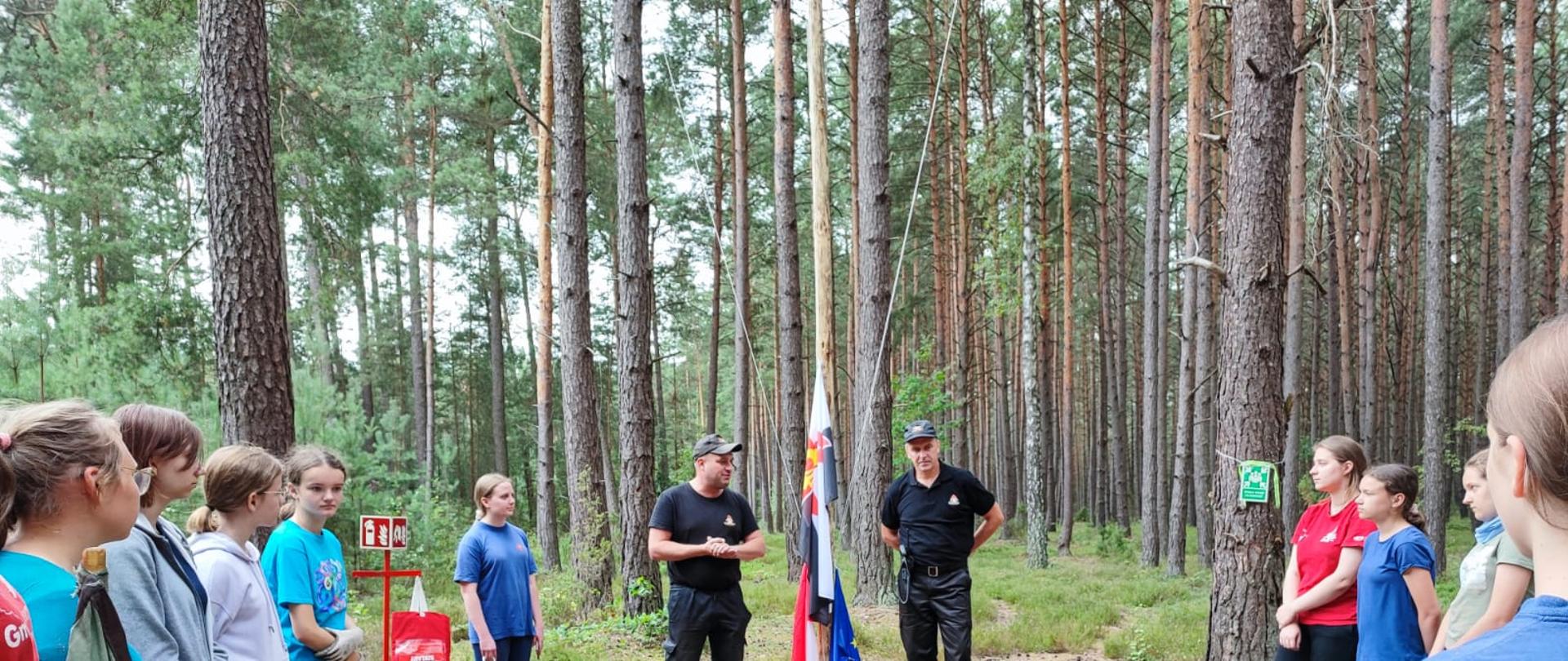 Na polanie w lesie stoi dwóch strażaków i grupa dziewcząt. Po lewej stronie obok dziewczynek stoi czerwony stojak na którym wisi koc gaśniczy. Przy stojaku stoi gaśnica. Dookoła polany rosną wysokie drzewa. 