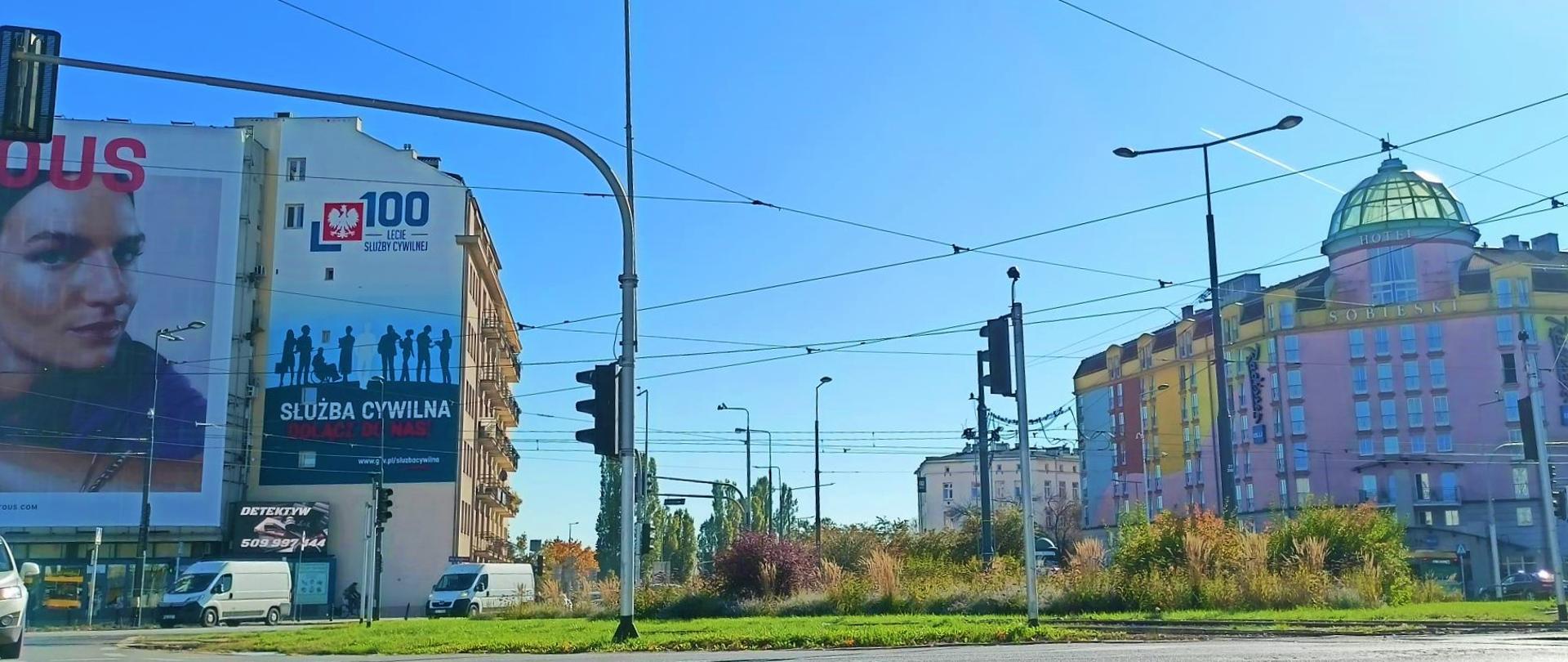 Plac Zawiszy w Warszawie. Na ścianie kamienicy mural rocznicowy, który przedstawia logo 100-lecia służby cywilnej, poniżej osiem granatowych zarysów postaci, a pod nimi biały napis "służba cywilna". 