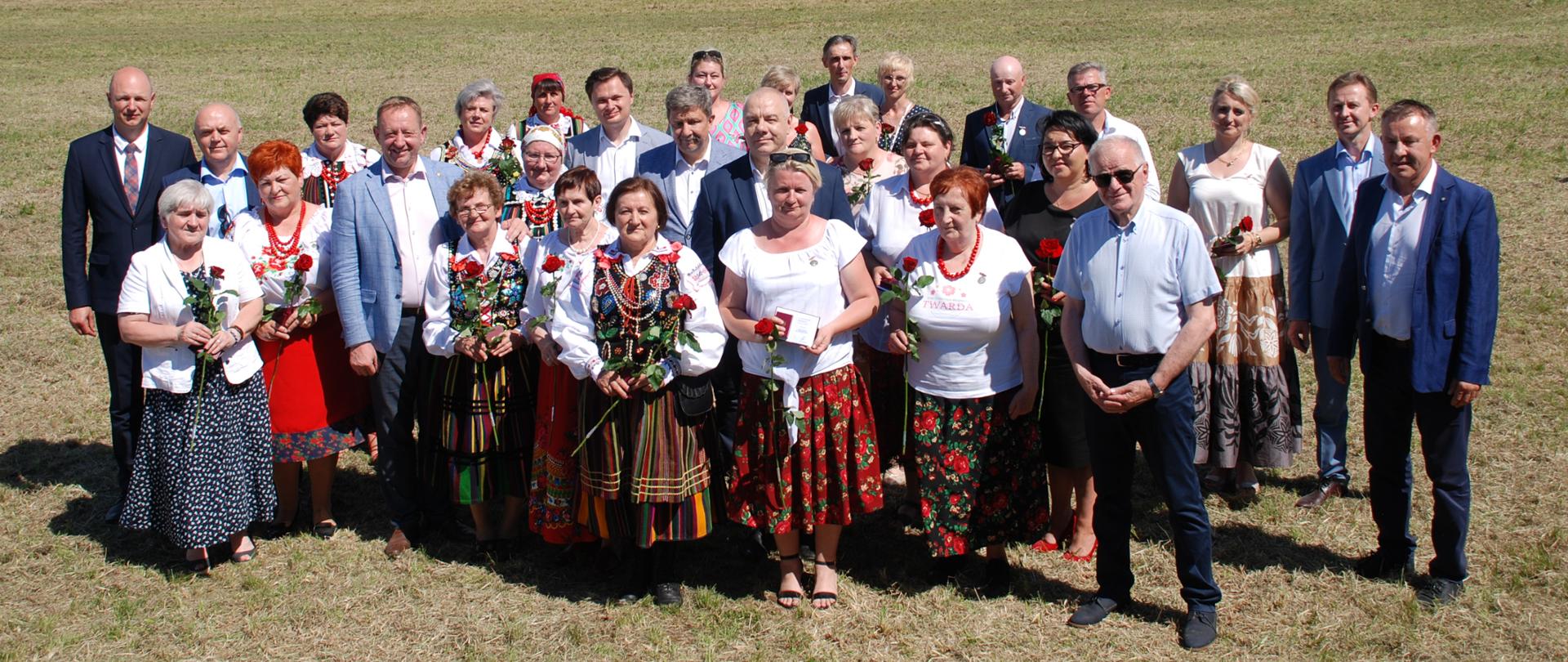 Na zdjęciu bardzo duża grupa osób, część Pań ubrana w regionalne stroje. Zebrani stoją na trawie, w tle rzeka i lasy. 