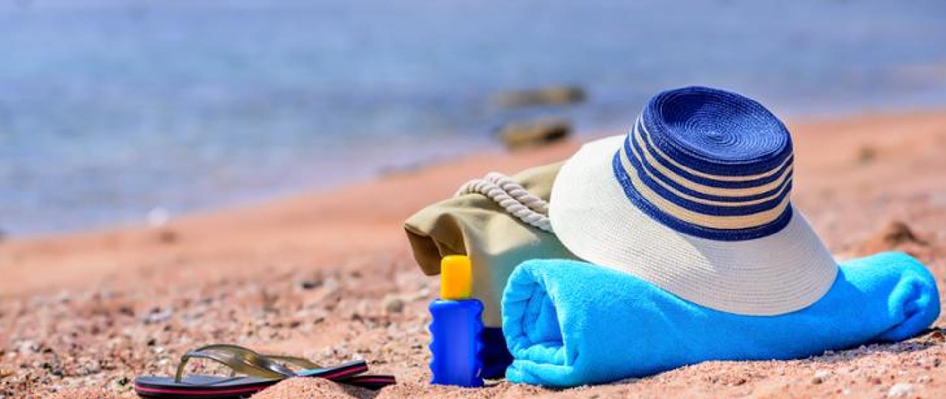 Na plaży leżą niebieski ręcznik, materiałowa torba. Ma nich leży słomkowy kapelusz, a obok widać klapki i olejek.