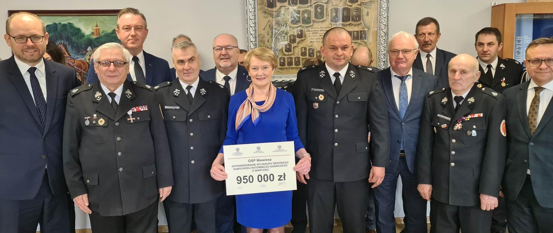 Grupowe zdjęcie przedstawicieli jednostki OSP posłów . Na środku stoi pani w niebieskim stroju i trzyma promese z napisem OSP niewiesz dofinansowanie do zakupu śreniego samochodu ratowniczo-gaśniczego o wartości 950 000 zł