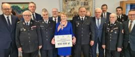 Grupowe zdjęcie przedstawicieli jednostki OSP posłów . Na środku stoi pani w niebieskim stroju i trzyma promese z napisem OSP niewiesz dofinansowanie do zakupu śreniego samochodu ratowniczo-gaśniczego o wartości 950 000 zł