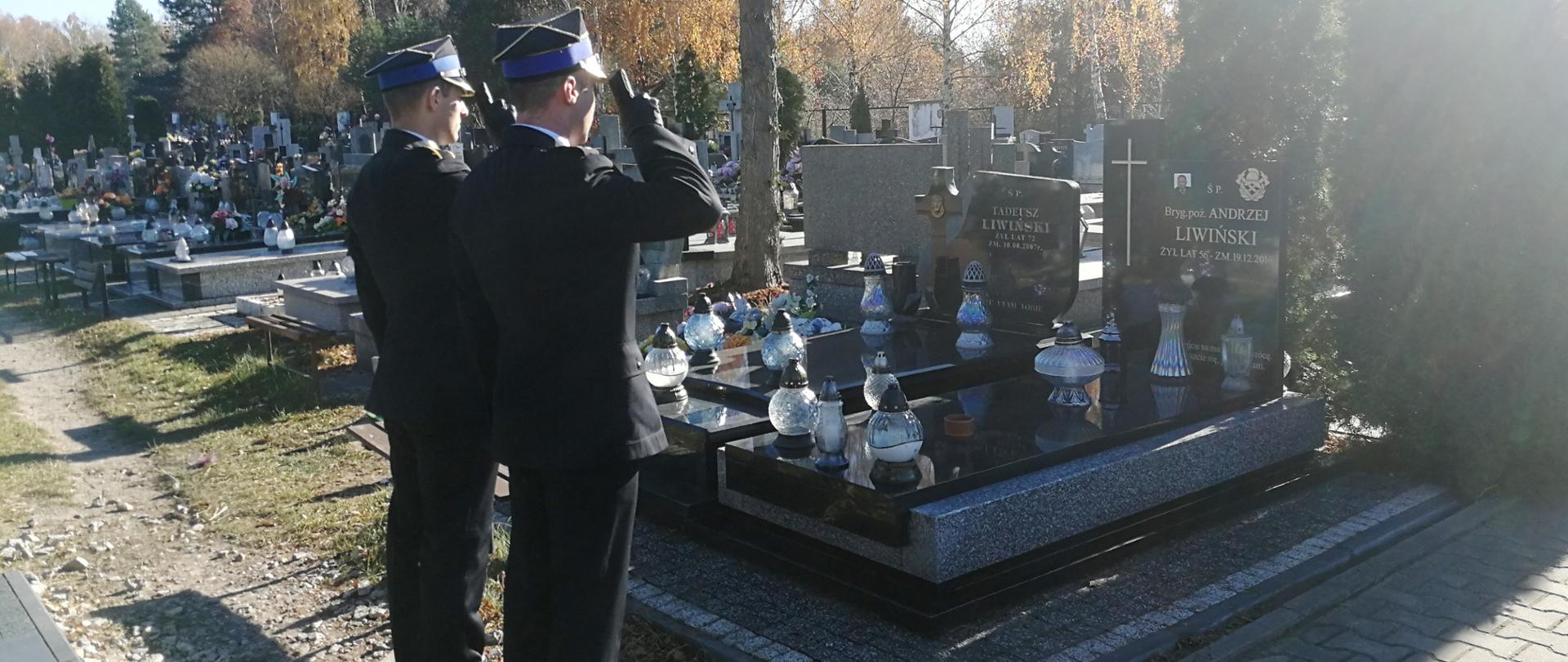 Na zdjęciu widać strażaków oddających hołd zmarłemu byłemu funkcjonariuszowi straży pożarnej. W tle widać inne pomniki znajdujące się na cmenatrzu oraz roślinność.