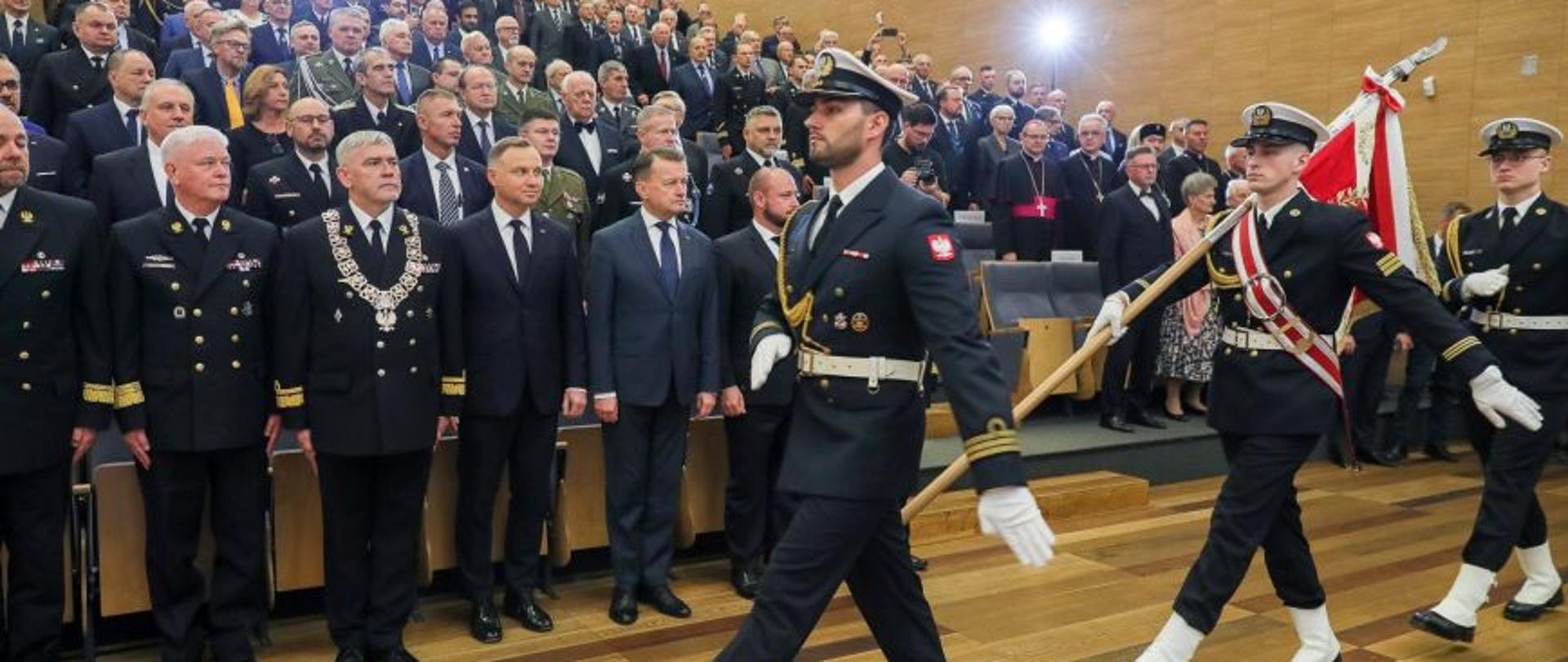 Poczet sztandarowy maszeruje przed zaproszonymi gośćmi wojskowymi osobami cywilnymi. 