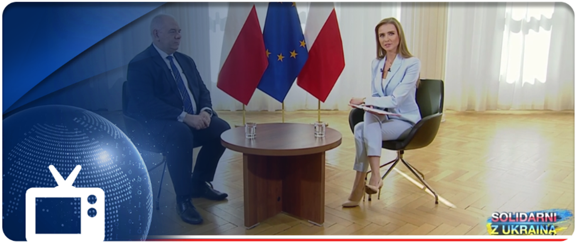 Z lewej strony wicepremier Jacek Sasin, z prawej strony redaktor Ewa Bugała, w środku stolik, w tle flagi. W lewym dolnym rogu ikonka telewizora. 