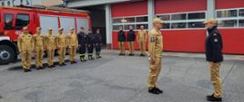 Na zdjęciu widać grupę strażaków stojących w szeregu przed remizą strażacką i czerwonym wozem pożarniczym. Dwóch strażaków znajduje się na pierwszym planie, zwróconych do siebie podczas oficjalnej zbiórki lub odprawy. Większość osób ma beżowe mundury, część ubrana jest w czarne stroje służbowe. W tle znajduje się budynek Komendy Powiatowej Państwowej Straży Pożarnej z czerwonymi bramami garażowymi. Niebo jest pochmurne, a plac przed budynkiem mokry.