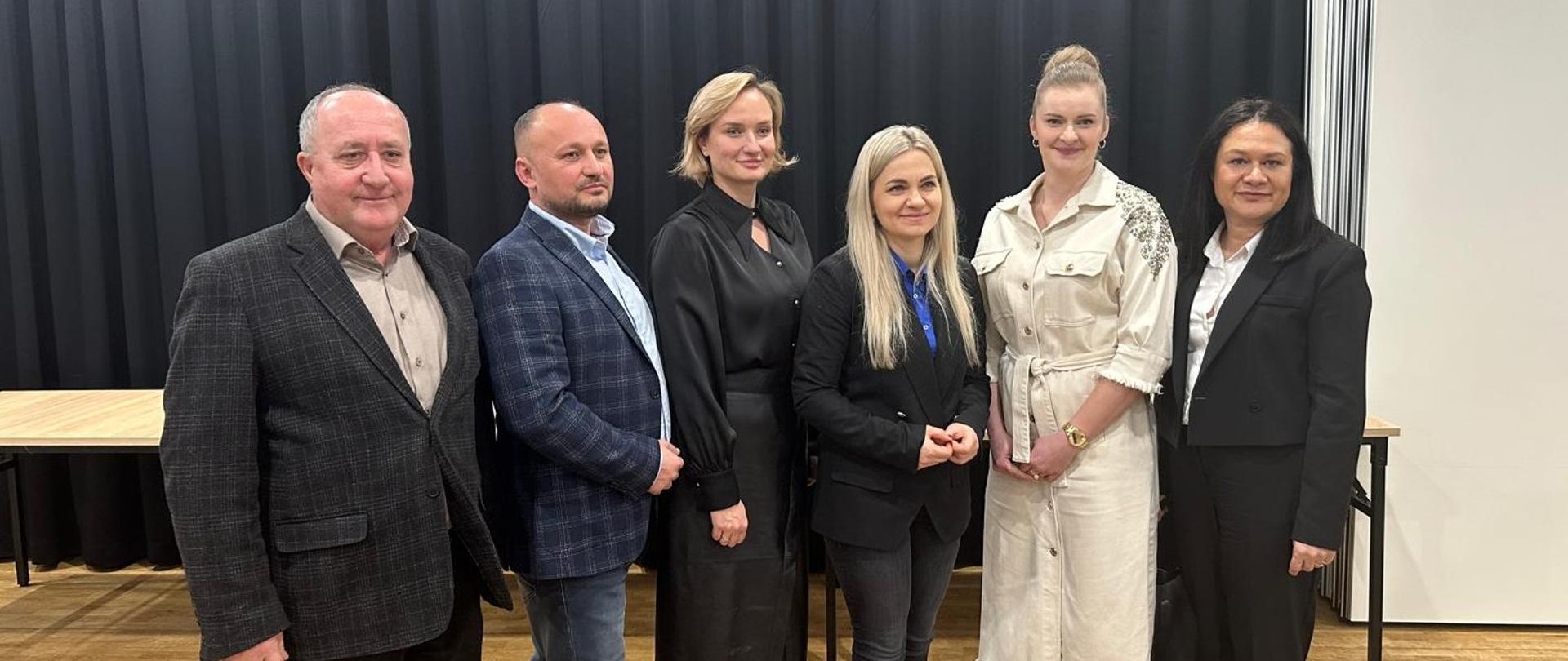 Siedem osób stojących obok siebie podczas oficjalnego spotkania, ubranych w stroje biznesowe, na tle ciemnej kurtyny w sali konferencyjnej