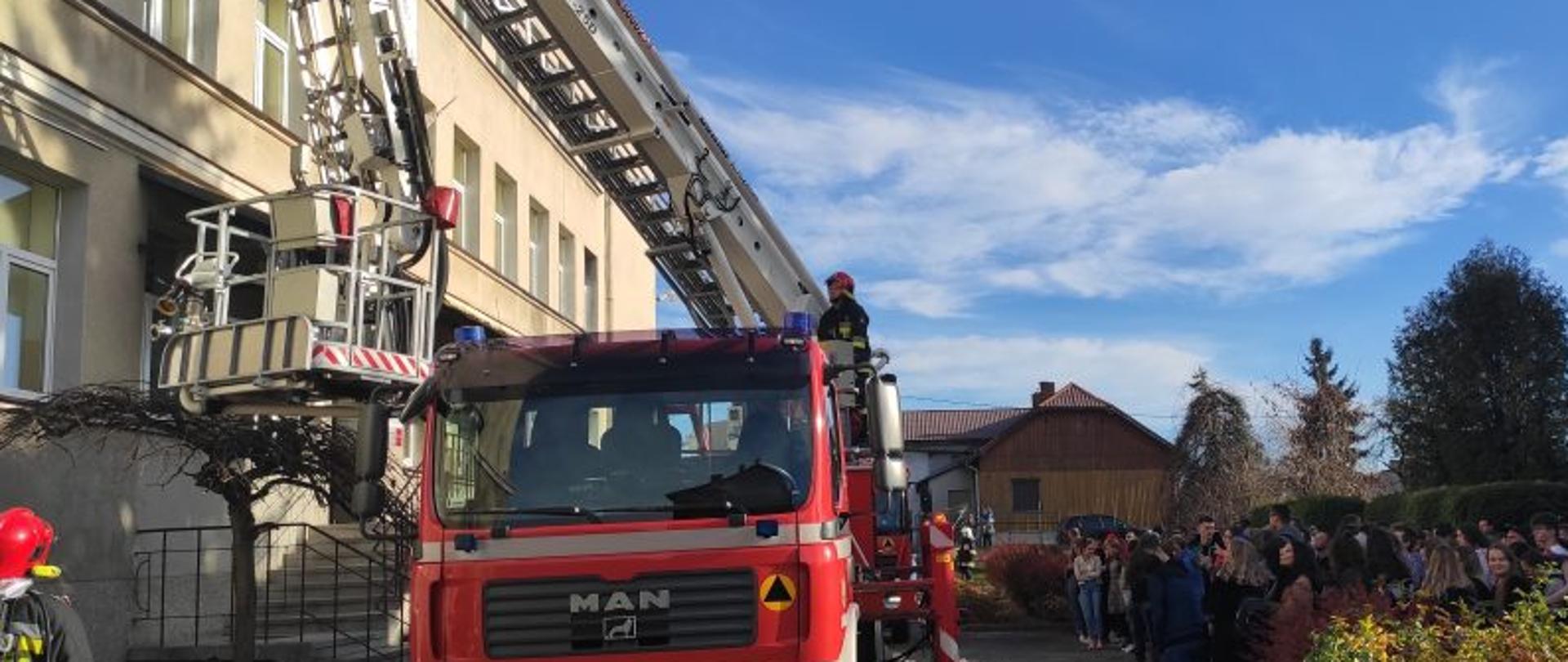 Na zdjęciu budynek szkoły, obok szkoły samochód pożarniczy z drabiną, na samochodzie strażak operator drabiny, obok samochodu uczniowie liceum, w tle dom drewniany, drzewa 