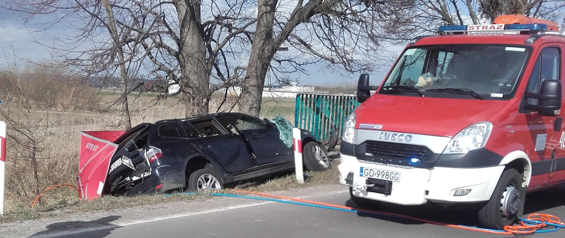 Samochód ratowniczy stoi na jezdni, rozbity pojazd znajduje się poza jezdnią