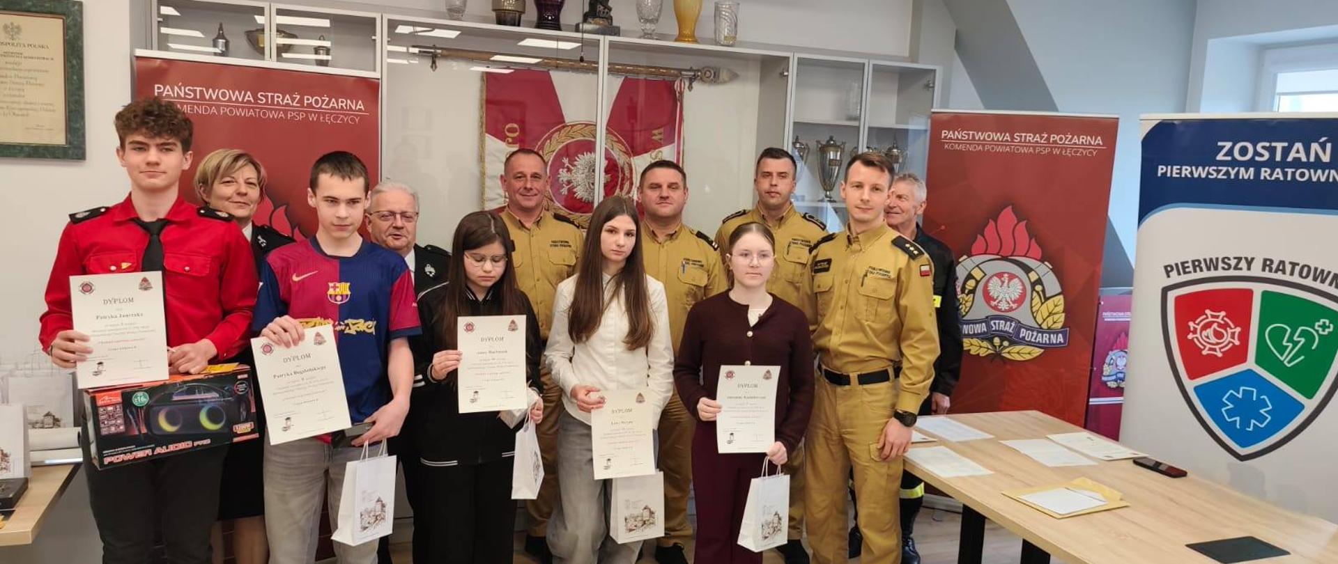 Na zdjęciu widoczna jest grupa ludzi - kilku strażaków w mundurach stoi razem z dziećmi, którzy trzymają dyplomy oraz nagrody. Wygląda to na uroczyste wręczenie wyróżnień po zakończeniu konkursu. W tle są gabloty z pucharami oraz banery Państwowej Straży Pożarnej. Wszyscy ustawieni są do wspólnego zdjęcia.