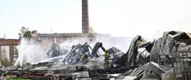 W centrum strażacy dogaszający pogorzelisko. Pracują w aparatach ochrony dróg oddechowych. Na zdjęciu linie gaśnicze, fragmenty konstrukcji spalonej hali, rozlewisko piany gaśniczej. W tle widoczny murowany z cegły budynek z wysokim kominem.