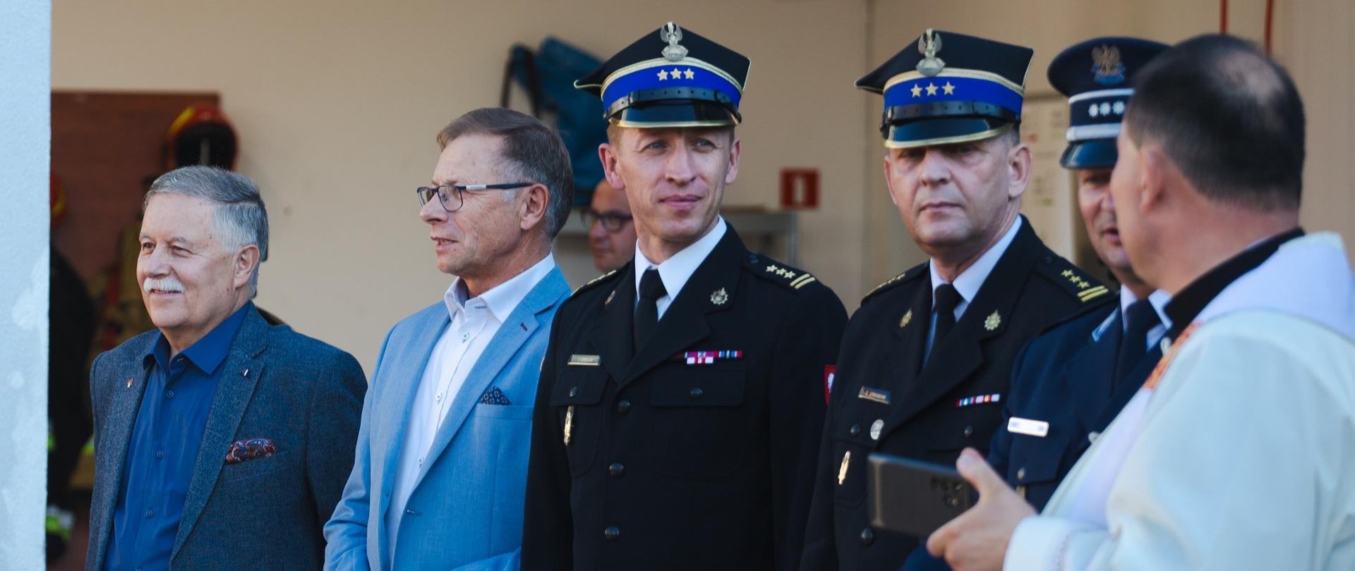 grupa mężczyzn , w tym strażacy w mundurach i rogatywkach stoją obok siebie, zwróceni w stronę księdza