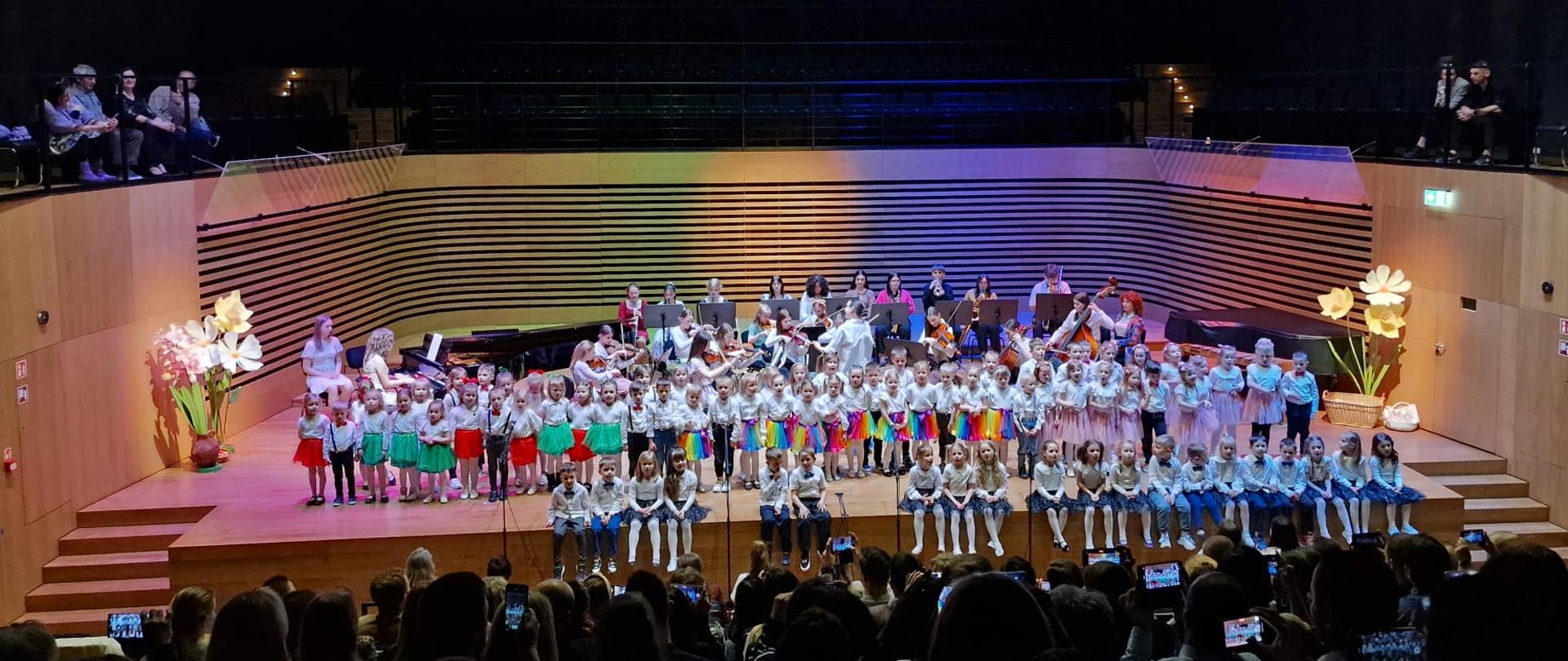 Dzieci stojąc lub siedząc w trzech rzędach śpiewają, za nimi kobieta dyryguje orkiestrą kameralną.